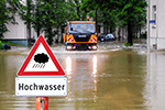 Hochwasserschild vor überfluteter Straße, Straßenmeisterfahrzeug im Hintergrund